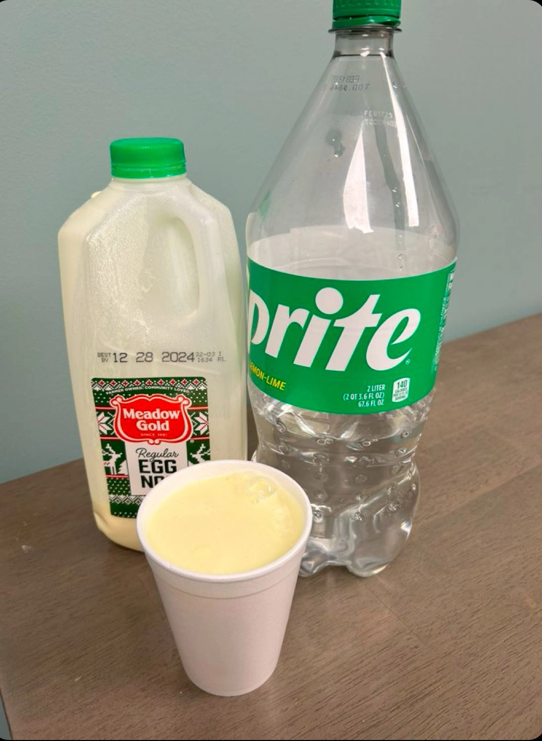 a bottle of egg nog next to a bottle of sprite, with an ominous cup of light yellow substance sitting between them like a terrible bastard child